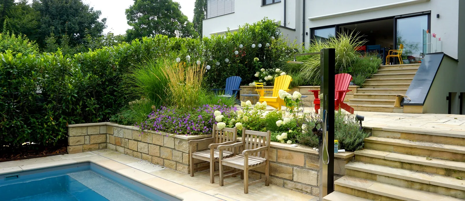 Garten mit Pool, Natursteinstufen und blühenden Stauden vor einem Wohnhaus