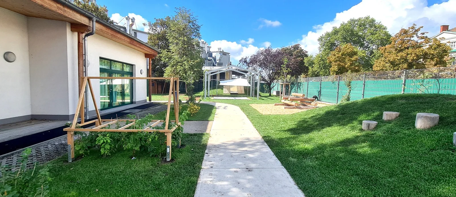 Garten eines Kindergartens mit Weg, Rasen, Spielhügel und Bäumen